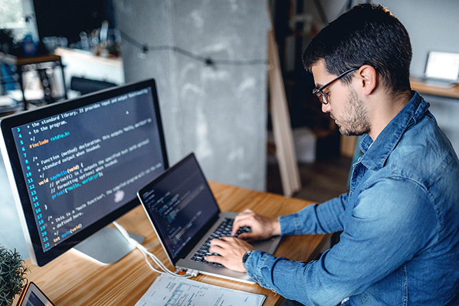 Photo of man coding on a laptop with a large computer monitor display