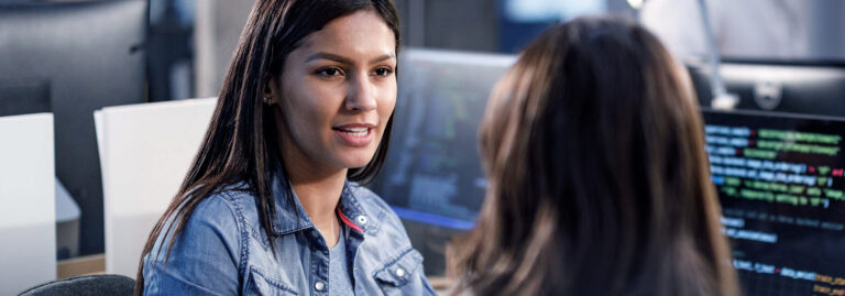 Two female tech colleagues talking at their desks in an office